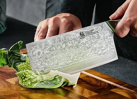 Image of man holding a huge Japanese cleaver knife from which he is cutting spinach on a wooden cutting board.