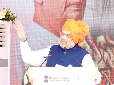 Image of HM Amit Shah wearing a orange turban and addressing a crowd on Hyderabad Liberation day. He has raised his right hand in the image while speaking to the audience present.