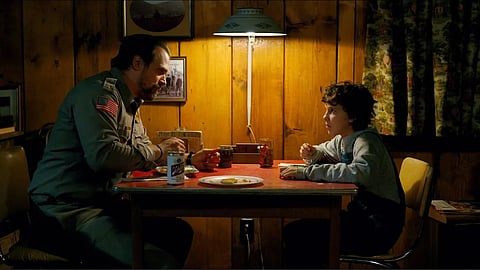 Millie Bobby Brown and David Harbour eating together at a dining table.