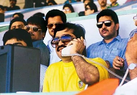 Image of Dawood Ibrahim watching a match. He is wearing a yellow t shirt with shades and he has got a white phone on his hand.