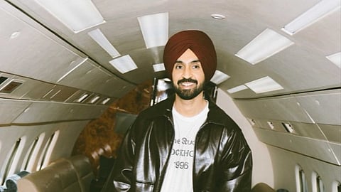 Diljit Dosanjh posing on his way to Australia for the Aura Tour 2025. He is wearing a black jacket and a maroon turban.