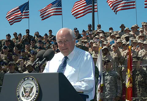 Image of Former US Vice President Dick Cheney addressing a crowd in front of a podium. He has got the US army in his background and 4 flags of the Unites States of America.