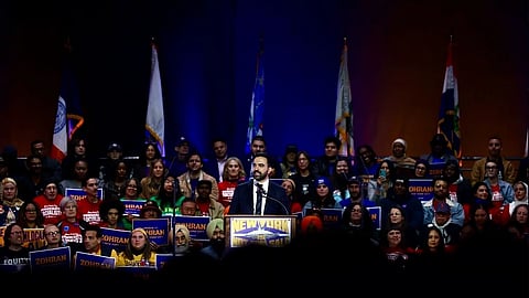 Zohran Mamdani speaks at a podium labeled "New York" surrounded by supporters holding signs. Diverse crowd with energetic and supportive expressions. Flags line the stage.