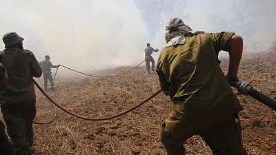 Firefighters work together to control fire, pulling a large hose across a dry, smoky field as thick smoke fills the air.