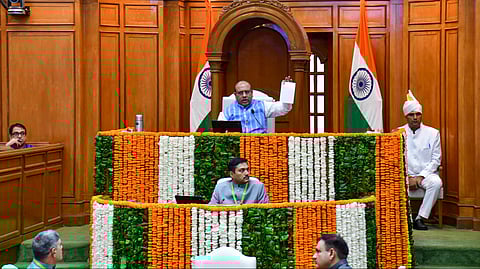 The speaker in the Delhi Assembly hall holds up a document while addressing members. The podium is decorated with orange, white, and green flower garlands, and two Indian national flags are displayed in the background.