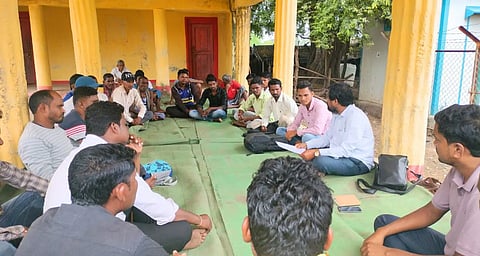 A gram panchayat sitting together on green mats in a verandah, discussing an issue.