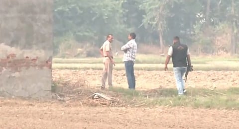 Three men stand in a field, engaged in conversation. One, wearing a uniform, faces a man in a check shirt, while another holds a camera. Trees are in the background.