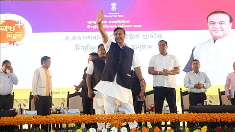 Chief Minister of Assam Hemanta Biswa Sarma in an event. He is standing on the stage raising his arm and smiling. There are people in the background of the party with a banner featuring the CM in the background.