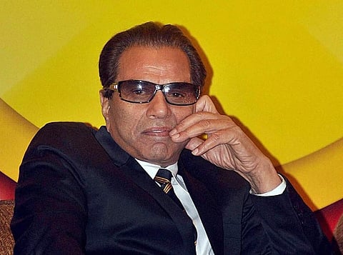 Image of Sholay actor Dharmendra wearing his classic suit with a stripped tie. He is wearing brown shades. His one hand is on his chin.
