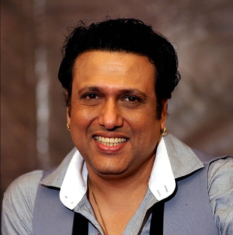 Actor Govinda Smiling with short dark hair, wearing a light gray shirt and a dark vest. The background is a soft, blurred brown, creating a warm atmosphere.