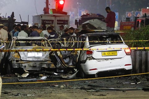 Two burnt cars are cordoned off by police tape at night near red Fort, surrounded by onlookers and emergency vehicles. A red traffic light glows in the background. the image depicts the scene of Read Fort Blast in New Delhi.