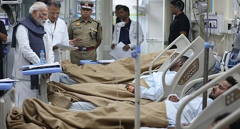 PM Modi visits a hospital ward, speaking with a doctor. Several patients lie in beds covered with brown blankets. The atmosphere is serious and compassionate.
