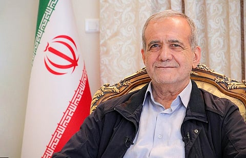 President Masud Pezeshkian sits on a decorative chair, smiling warmly. Behind him is an Iranian flag. The atmosphere is formal yet approachable, with soft lighting.