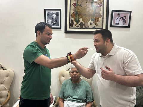 Tej Pratap Yadav and Tejashwi Yadav stand in a room, one feeding a sweet to the other with a spoon, while lalu Yadav sits between them. Family photos and art are on the wall.