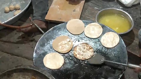 Several round Sattu Paratha cooking on a large griddle, with a person flipping them using a spatula. Nearby, a bowl of yellow liquid, likely ghee, adds a warm tone.