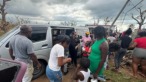 A group of people gathers around a van distributing supplies in a disaster-stricken area. The scene is intense, with cloudy skies and damaged trees.