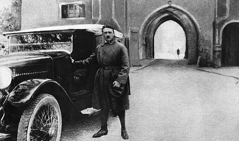 Black and white Image of Adolf Hitler standing beside his car. In the background a pathway is seen from where a shadow of one person can be seen. Hitler and his car is positioned at the left side of the image.