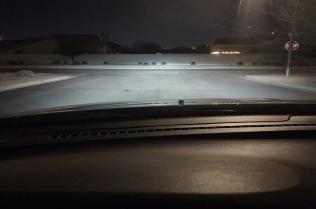 View from a car dashboard at night, headlights illuminating a residential street. A stop sign is visible on the right. The mood is calm and quiet.