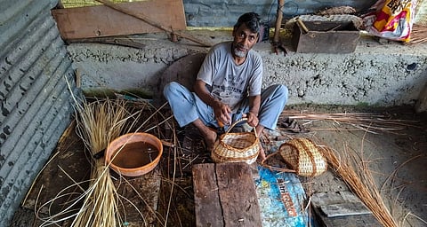 A man sits on the floor, weaving baskets out of willow branches. He is wearing a t shirt and jeans and has a grey beard. The floor is mud.