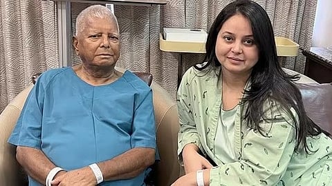 Lalu Prasad Yadav and a Rohini Acharya sit together in a hospital room. The man wears a blue hospital gown, looking calm. The woman, in a patterned gown, smiles softly.
