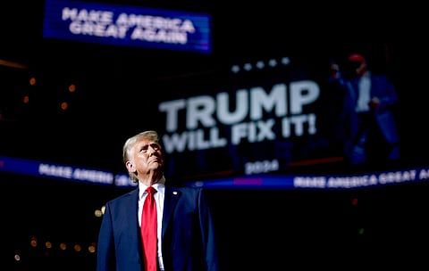 Donald Trump in a dark suit and red tie stands on stage, looking upward. Behind him, a screen displays "Trump will fix it!" with a slogan. The atmosphere is dramatic.