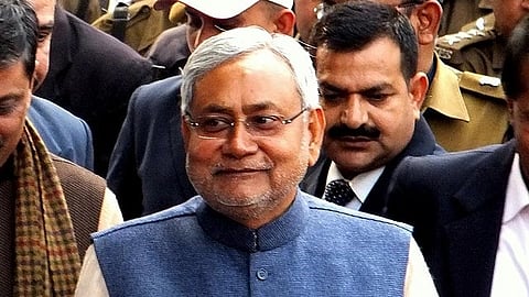 Nitish Kumar smiling man in a blue Nehru jacket stands among a group of people, some in uniform, suggesting a formal or official event. The mood appears positive.