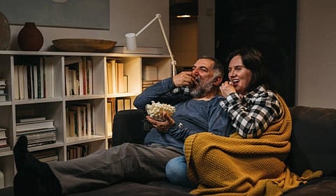 A couple sits on a cozy couch watching TV, sharing a bowl of popcorn and wrapped in a yellow blanket. They appear relaxed and amused.