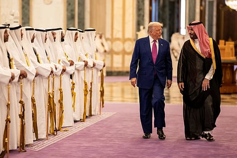 Donald Trump in a blue suit and pink tie walks beside Prince Mohammed bin Salman in traditional Middle Eastern attire on a purple carpet, flanked by a ceremonial guard. The setting appears formal and regal.