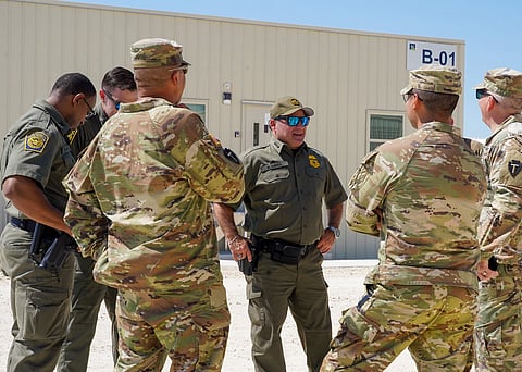 A group of uniformed military and law enforcement personnel in a discussion outdoors. The setting is sunny, near a beige building labeled "B-01." The tone is serious.
