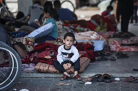 A young boy sits on a thin mattress among scattered belongings, blankets, and other displaced families sheltering in a public space in Gaza following heavy airstrikes