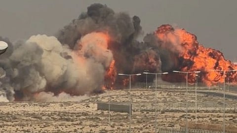 Large fire and thick smoke rise from a desert area, with intense orange flames. In the foreground, several airport service vehicles are parked. The scene conveys urgency and danger.