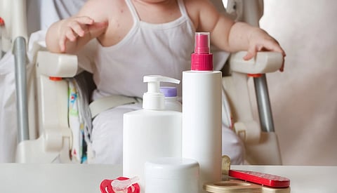 A baby in a white outfit sits in a high chair, with various baby care products like bottles and a pacifier on the table in the foreground. The scene conveys a clean, nurturing environment.