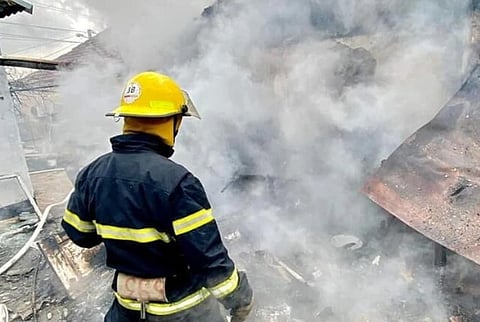 A Ukrainian firefighter in a yellow helmet and dark uniform surveys a smoky scene, with thick gray smoke billowing around. The atmosphere is tense and urgent.