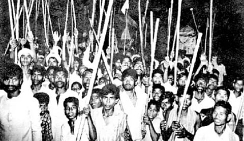 A black-and-white photo of a large group of people, holding sticks, raising them in a unified gesture. The mood suggests solidarity and determination.