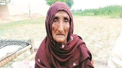 Elderly woman in a maroon headscarf sitting outdoors on a woven cot, surrounded by grass and trees. Her expression is calm and reflective.