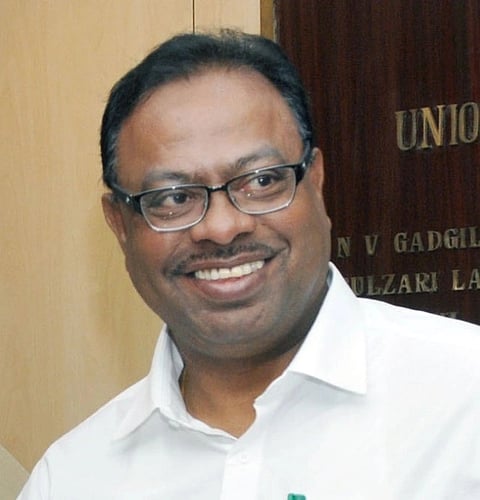 Chandrashekhar Bawankule wearing glasses and a white shirt smiles warmly, standing against a wooden background with engraved text partially visible. The mood is friendly.