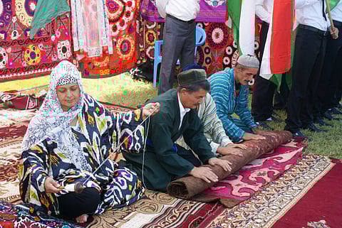 A Tajik woman and 2 men siiting on a carpet. The woman is wearing a headscarf and is unravelling thread from a spool. The men are wearing caps and rolling up a carpet. They are dressed in traditional vibrant Tajik dress.