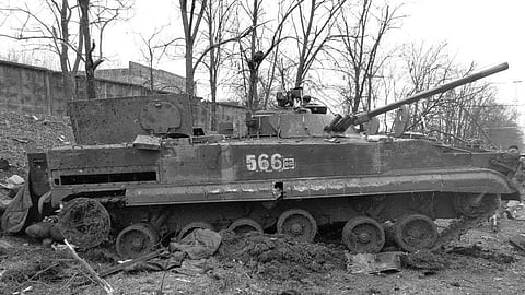 A damaged military tank sits abandoned in a muddy, debris-filled environment. Leafless trees and a concrete wall appear in the background, conveying desolation.