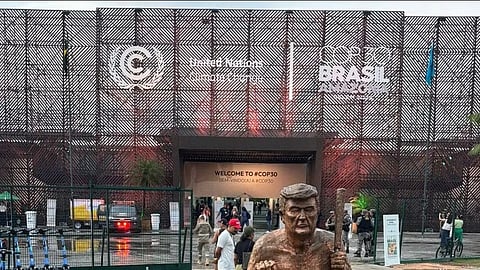 A bronze statue of a child carrying an overweight man on his shoulders stands on grass. Behind is the COP30 venue entrance with people and signs visible.