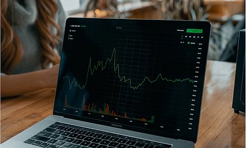 Laptop displaying a stock market graph with fluctuating green line on a grid, set on a wooden table. A person is partially visible, creating a focused atmosphere.