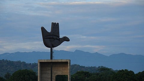 A large open hand sculpture stands on a concrete base in a grassy park. People casually walk nearby. Mountains and a cloudy sky form the backdrop.