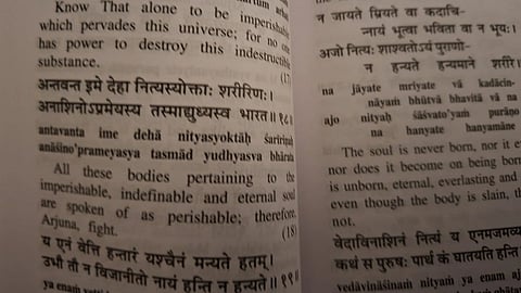 Close-up of an open book showing text in Sanskrit and English. The passage discusses themes of the soul's imperishability and eternal nature, from the Bhagavad Gita. The tone is philosophical and introspective.