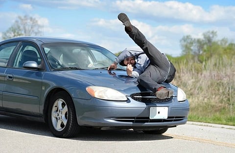 A man in a gray suit is falling onto a blue car's hood on a rural road, capturing a moment of surprise and urgency under a bright blue sky.