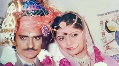 A couple in traditional Indian wedding attire, both adorned with pink flower garlands. The groom wears a colorful turban, and the bride has a red bindi, smiling gently.