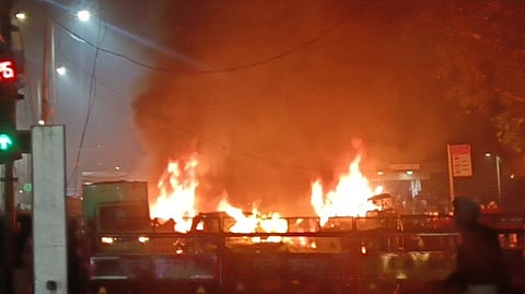 Street scene with large flames and smoke rising into the night sky. Silhouettes of barriers and people in the foreground create a tense atmosphere.