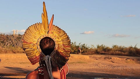 A person wears a large, ornate feather headdress and red bead necklace, pointing across a sunlit, arid landscape with scattered pools and sparse vegetation.