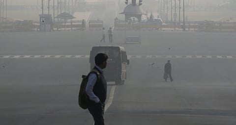 A person with a backpack walks across a wide, foggy road. A bus and a few people are visible in the background, obscured by thick haze, conveying pollution.