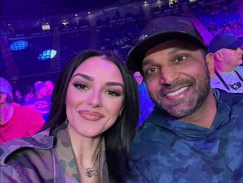 Image of FBI director Kash patel with his girlfriend Alexis Wilkins. in the image they are seen smiling at the camera in a lively indoor arena, illuminated by purple lighting. The background is filled with seated spectators.