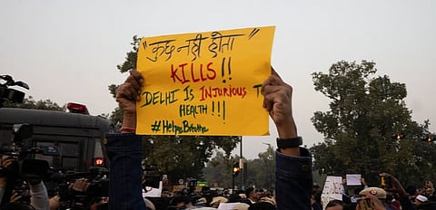 Protest scene with a person holding a yellow sign reading, "Delhi is injurious to health! #HelpUsBreathe," amidst a crowd and media cameras.