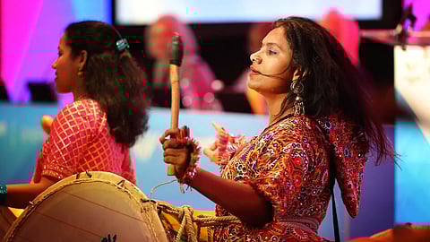 Two women in vibrant, patterned garments play drums energetically on a colorful stage, conveying joy and cultural celebration. Bright lighting enhances the scene.